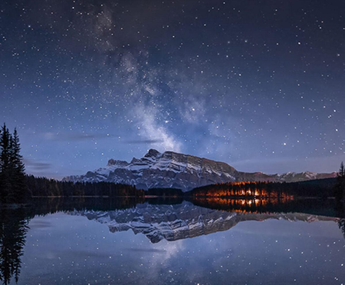 A panoramic nightscape featuring the Milky Way stretching across a still lake, with stars shimmering above a pine forest and snow-covered mountain.
