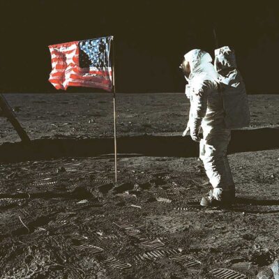 An astronaut stands beside the American flag on the surface of the Moon with visible boot prints in the lunar dust beneath a black space backdrop