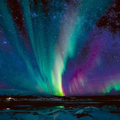 A vibrant aurora borealis in shades of green turquoise and purple dancing above a snow covered landscape and icy lake under a star filled sky