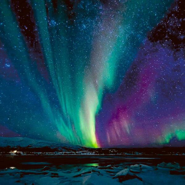A vibrant aurora borealis in shades of green, turquoise, and purple dancing above a snow-covered landscape and icy lake under a star-filled sky