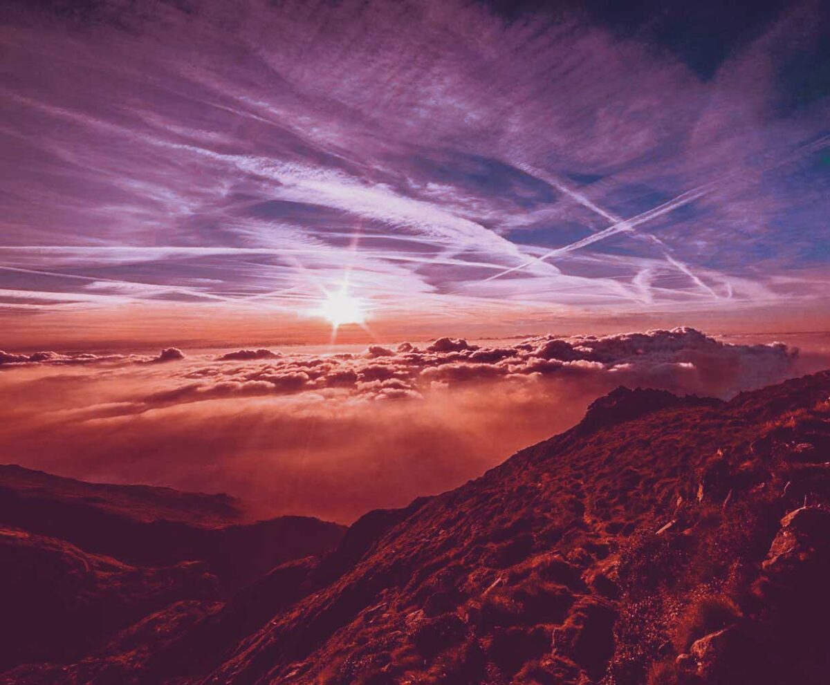 A sunrise emerging above the clouds over a mountain ridge, casting warm pink and orange hues across the sky