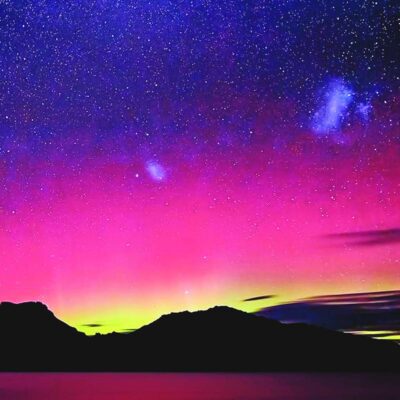 A surreal night sky filled with pink purple and electric blue hues from the aurora borealis above a dark mountain range and still lake