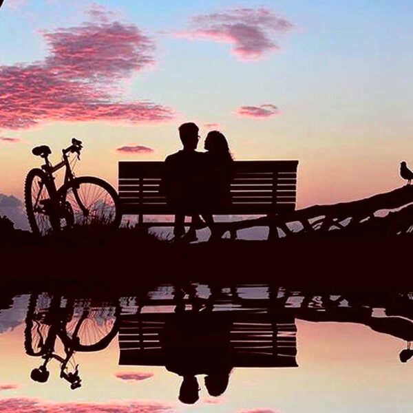 A couple sitting on a bench at sunset with a bicycle nearby, reflected perfectly in still water
