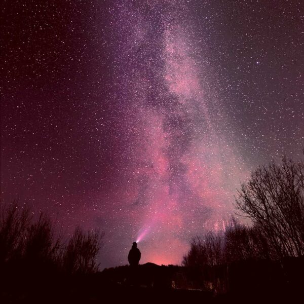 A person stands silhouetted against a starry sky with the northern lights in vibrant pinks and purples, casting a magical glow over the landscape."