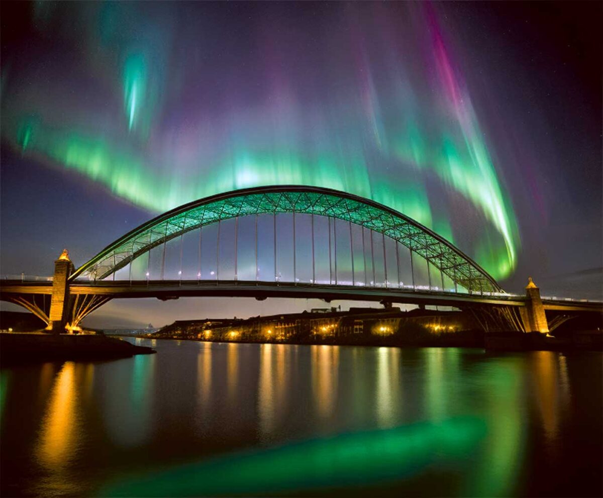 A vibrant display of the northern lights illuminates the sky above the iconic Newcastle bridge, reflecting in the calm waters below, with colors of green, pink, and purple swirling across the night sky.