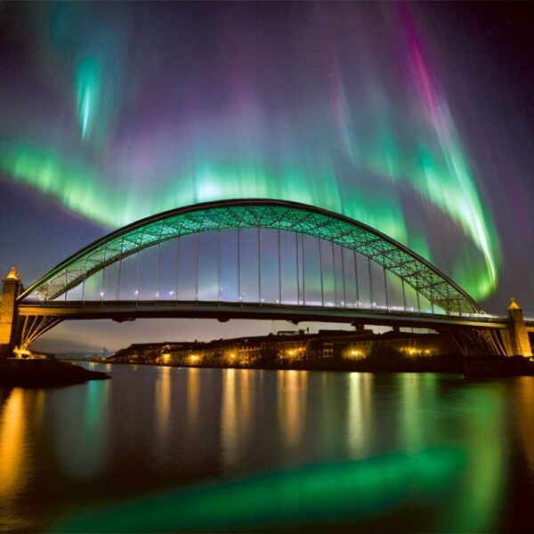 A vibrant display of the northern lights illuminates the sky above the iconic Newcastle bridge, reflecting in the calm waters below, with colors of green, pink, and purple swirling across the night sky.