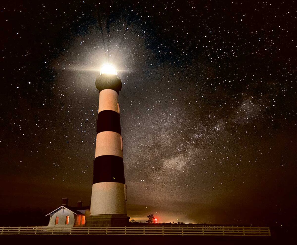A tall black-and-white striped lighthouse glowing at night under a star-filled sky and the Milky Way