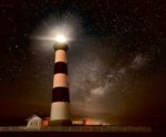 A tall black-and-white striped lighthouse glowing at night under a star-filled sky and the Milky Way