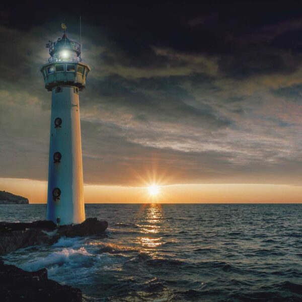 A lighthouse glowing at twilight with the sun setting over the ocean horizon and waves gently crashing below